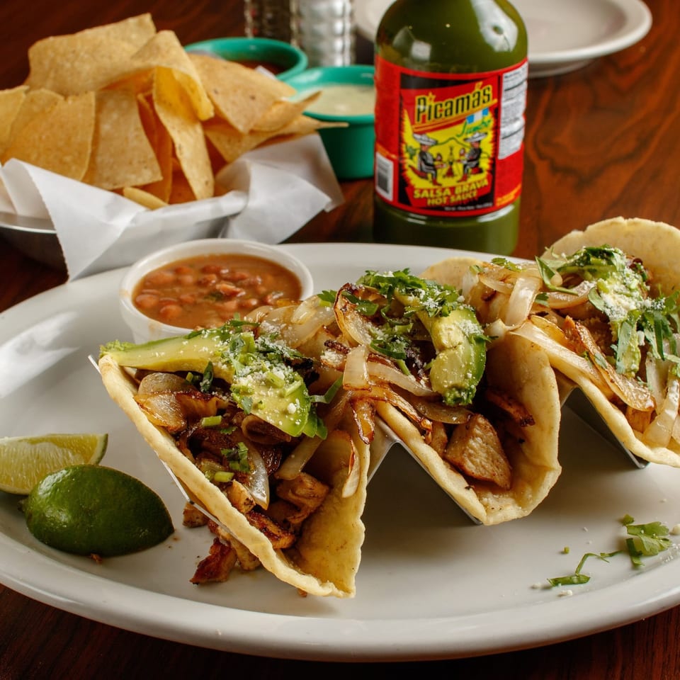 Three tacos with meat, onions, avocado, and cilantro, served with beans, chips, and hot sauce.