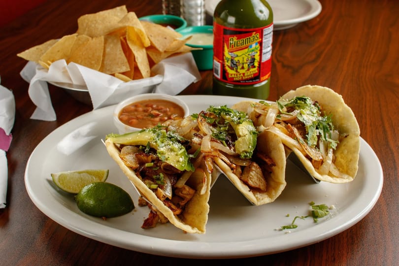 Three tacos with meat, onions, avocado, and cilantro, served with beans, chips, and hot sauce.