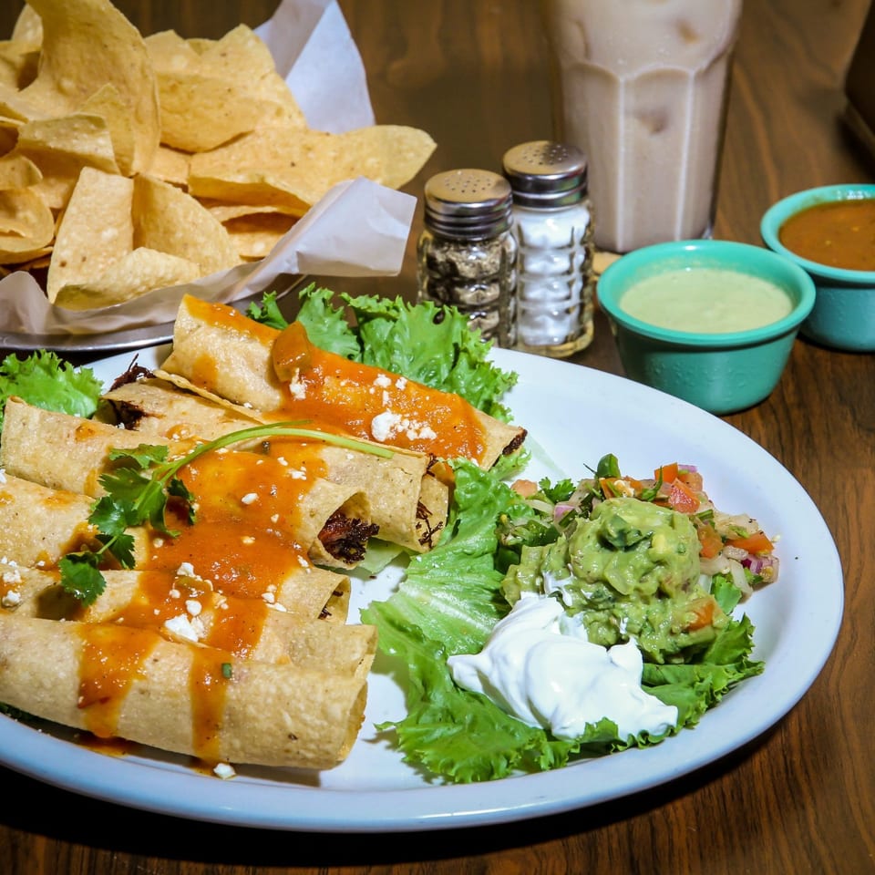 Mexican taquitos with red sauce, guacamole, sour cream, and lettuce, alongside chips and salsa.