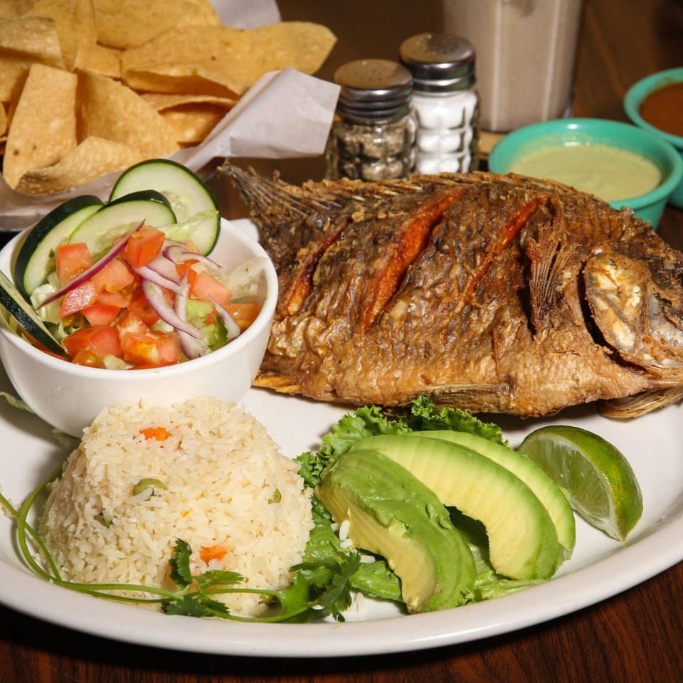 A whole fried fish with rice, salad, avocado, and lime, served with chips and salsa.