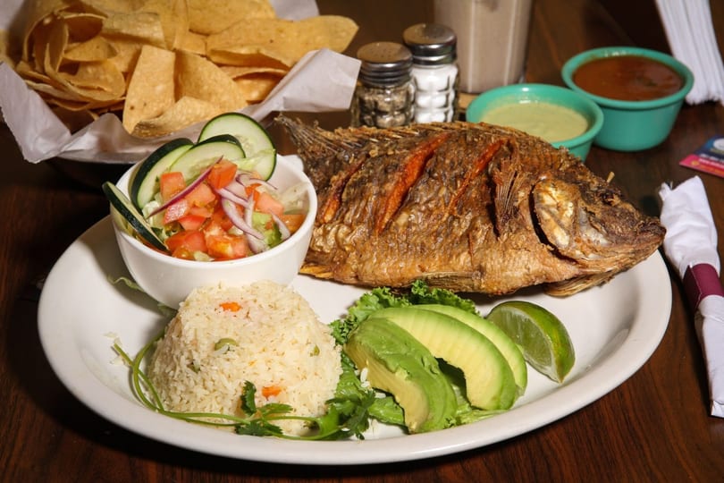 A whole fried fish with rice, salad, avocado, and lime, served with chips and salsa.