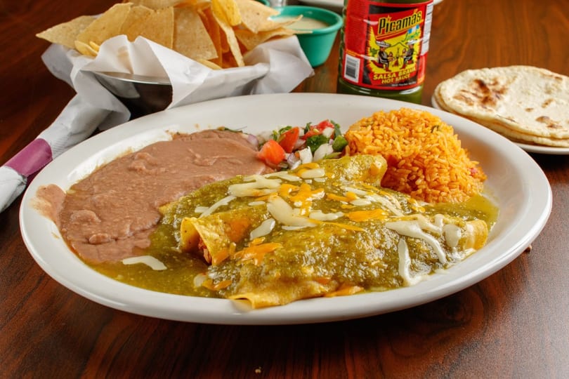 A plate of green enchiladas with rice, refried beans, pico de gallo, chips, and hot sauce.