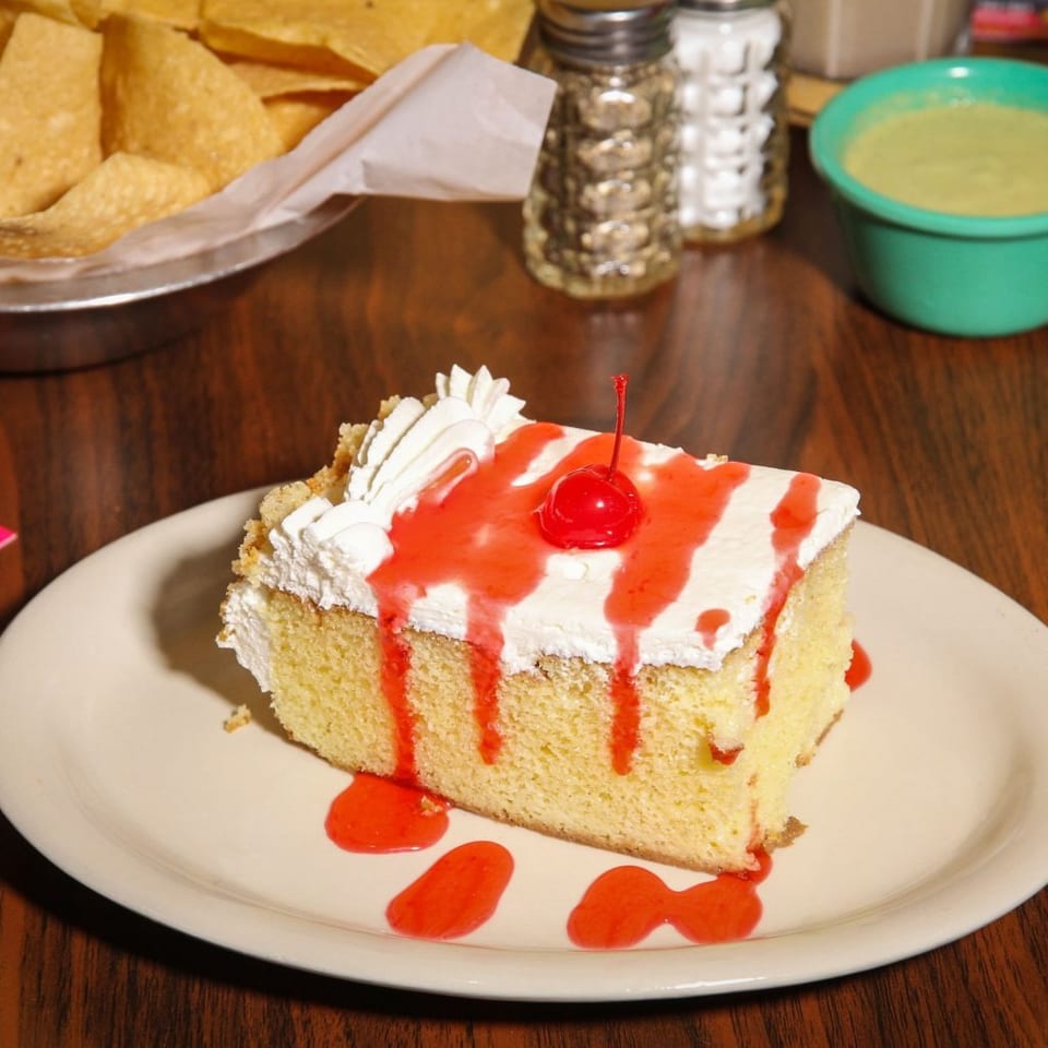 Slice of tres leches cake with whipped cream, strawberry drizzle, and a cherry on a white plate.