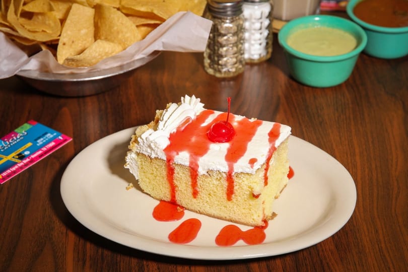 Slice of tres leches cake with whipped cream, strawberry drizzle, and a cherry on a white plate.