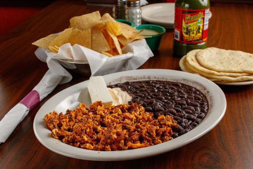 A plate of scrambled eggs with chorizo, black beans, cheese, and sour cream, with chips and tortillas.