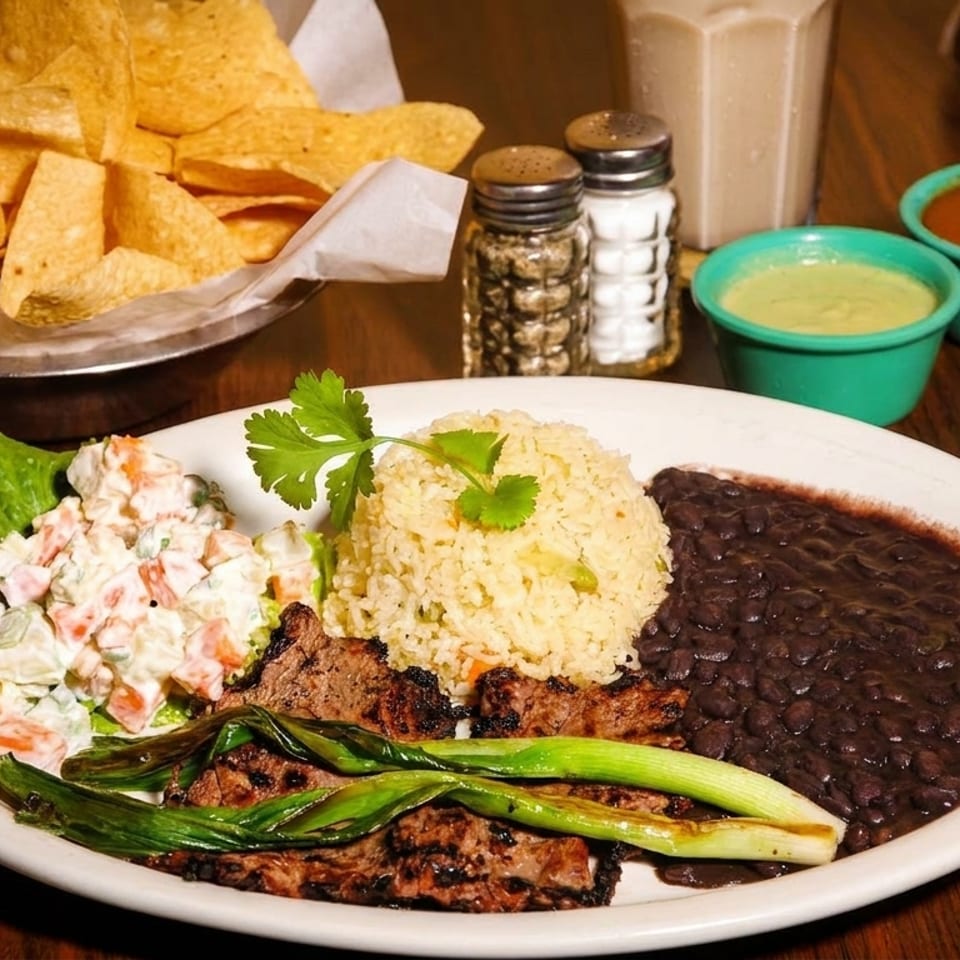 Grilled steak with rice, black beans, potato salad, and scallions, served with chips and salsa.