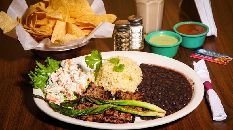 Grilled steak with rice, black beans, potato salad, and scallions, served with chips and salsa.
