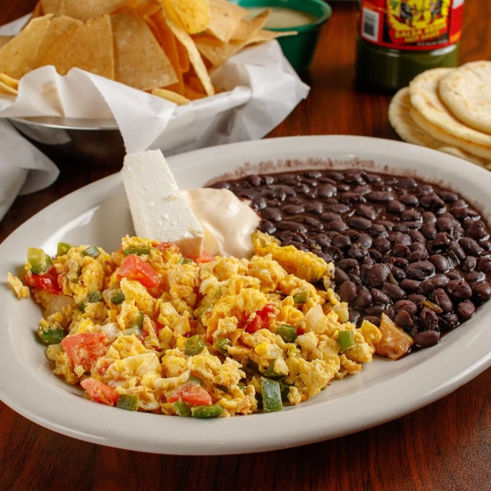 A plate of scrambled eggs with vegetables, black beans, cheese, and tortillas on a wooden table.
