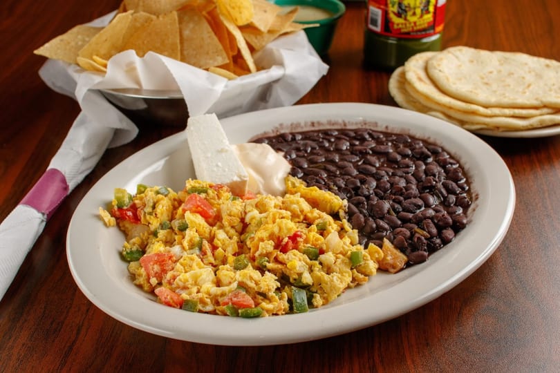 A plate of scrambled eggs with vegetables, black beans, cheese, and tortillas on a wooden table.