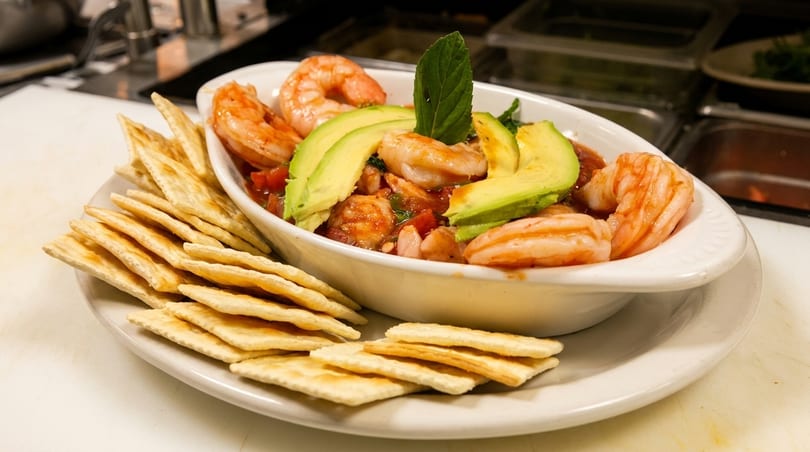 Shrimp ceviche with avocado slices and a mint garnish, served with crackers on a white plate.